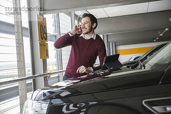 Lächelnder junger Mann  der mit seinem Smartphone spricht  während er sich auf die Motorhaube eines Autos auf einem Parkplatz stützt