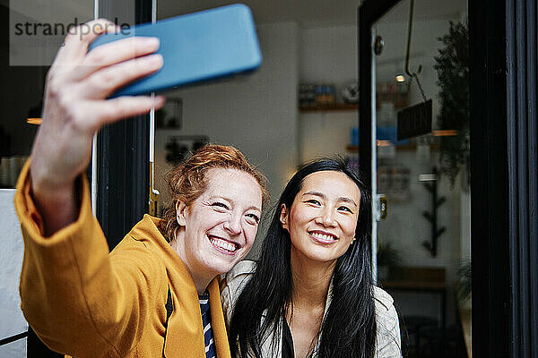 Lächelnde Frau nimmt Selfie durch Handy mit weiblichen Freund außerhalb Cafe