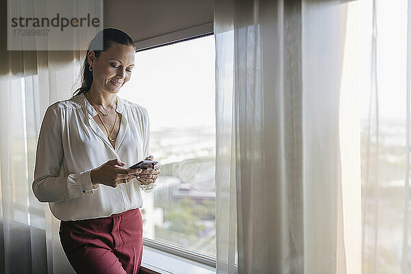 Weibliche Berufstätige  die mit ihrem Mobiltelefon SMS schreiben  während sie im Büro am Fenster stehen