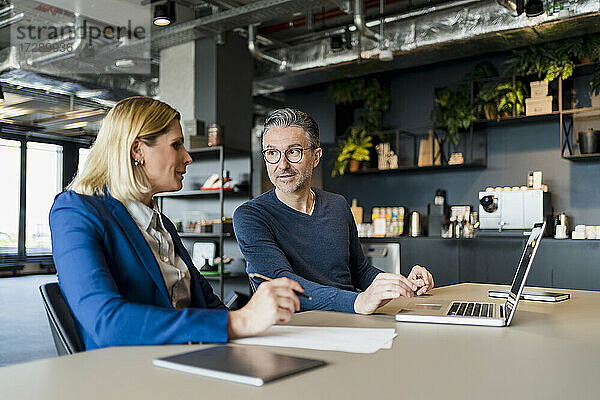 Männlicher Unternehmer im Gespräch mit einer weiblichen Fachkraft  die mit einem Laptop in einem kreativen Büro sitzt
