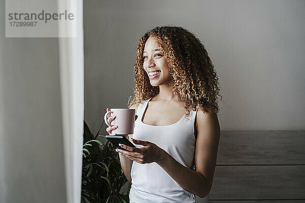 Glückliche Frau mit Smartphone und Kaffeetasse im Schlafzimmer stehend