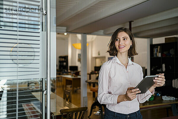 Lächelnde junge Frau mit digitalem Tablet  die im Büro steht und wegschaut