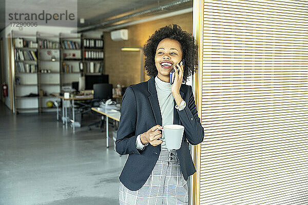 Junge Geschäftsfrau  die eine Kaffeetasse hält  während sie mit einem Smartphone im Büro telefoniert