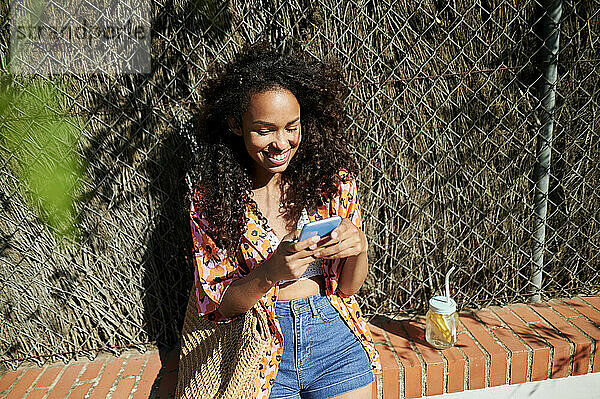 Lächelnde Frau  die ein Smartphone benutzt  während sie auf einer Stützmauer sitzt