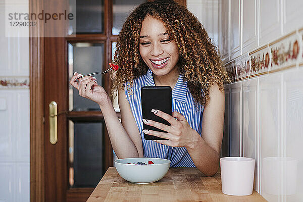 Lächelnde junge Frau benutzt ihr Smartphone beim Frühstück in der Küche