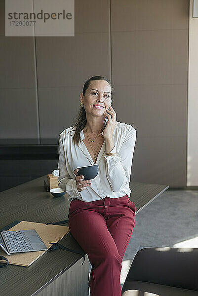 Lächelnde Unternehmerin  die eine Kaffeetasse hält und mit einem Smartphone spricht  während sie am Schreibtisch im Büro sitzt