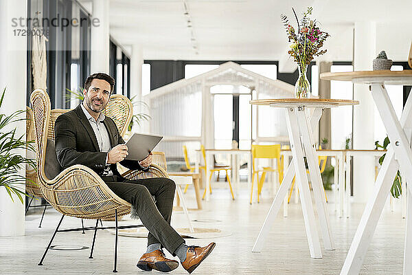 Lächelnder männlicher Unternehmer mit digitalem Tablet  der auf einem Stuhl in einer Büro-Cafeteria sitzt