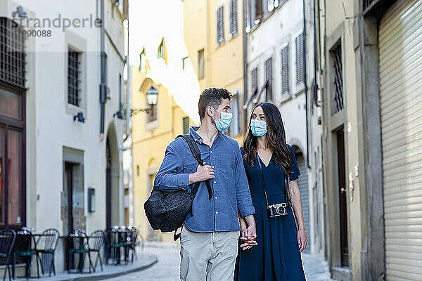 Touristenpaar mit Gesichtsschutzmaske  das sich auf einer Straße in der Stadt an den Händen hält