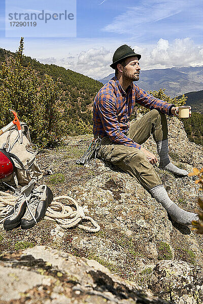 Männlicher Bergsteiger hält eine Tasse  während er an einem sonnigen Tag auf einem Berg sitzt