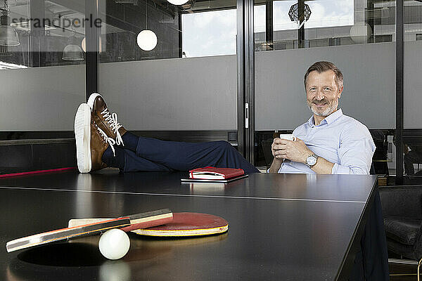 Lächelnder Geschäftsmann  der Kaffee trinkt  während er an der Tischtennisplatte im Büro sitzt