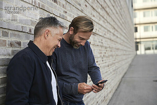 Fröhlicher männlicher Berufstätiger  der seinen Freund bei der Benutzung eines Smartphones an der Wand beobachtet