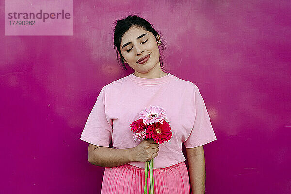 Lächelnde junge Frau mit geschlossenen Augen hält rosa Blumen vor einer Wand