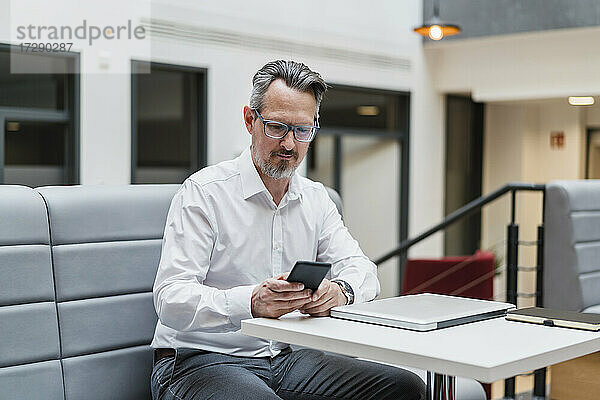 Männlicher Unternehmer  der ein Mobiltelefon benutzt  während er am Tisch im Büro sitzt