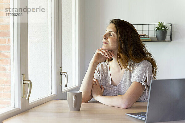 Frau mit geschlossenen Augen sitzt in der Nähe von Fenster zu Hause Büro
