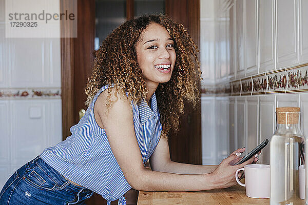 Lächelnde schöne Frau  die ihr Smartphone hält und sich auf den Tisch in der Küche stützt