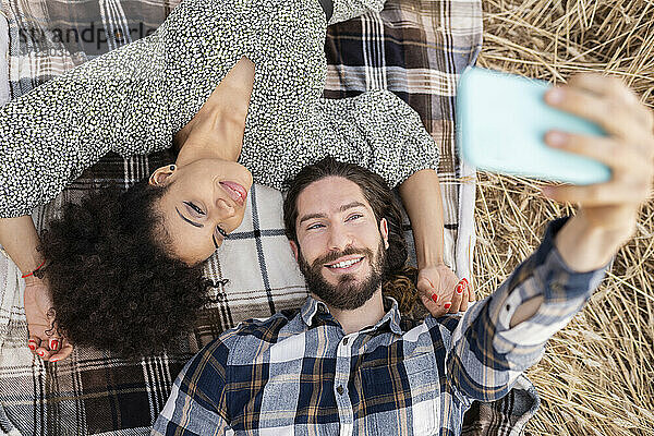 Lächelnder Mann  der ein Selfie mit einer Frau durch ein Mobiltelefon macht  während er auf einer Decke liegt