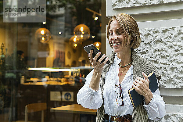 Lächelnde Frau  die ein Mobiltelefon benutzt  während sie vor einer Wand steht