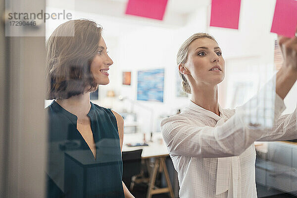 Lächelnde Geschäftsfrau  die eine Kollegin bei der Arbeit im Büro betrachtet  scheint durch Glas zu schauen