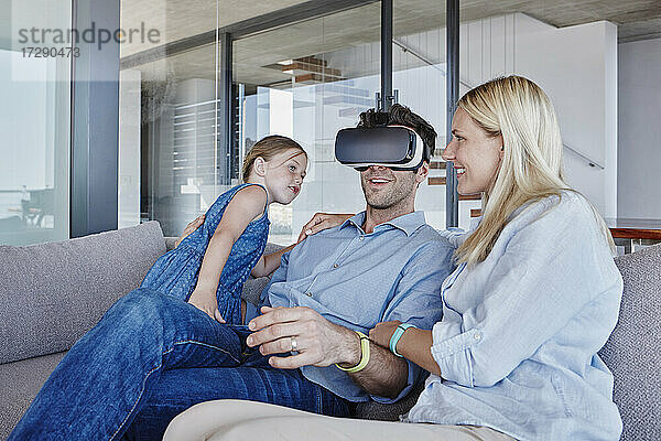 Lächelnder Mann  der einen Virtual-Reality-Simulator trägt  sitzt mit Frau und Tochter im Wohnzimmer