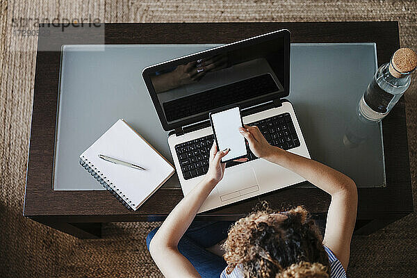 Geschäftsfrau  die zu Hause ein Mobiltelefon über einem Laptop benutzt