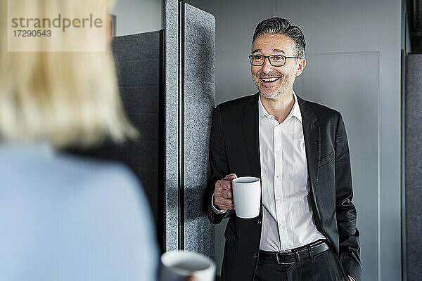 Lächelnder männlicher Unternehmer  der eine Kaffeetasse hält  während er mit einer weiblichen Fachkraft in der Pause im Büro spricht