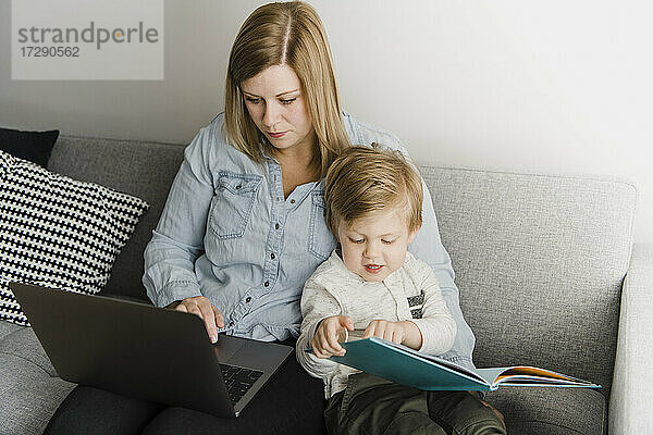 Mutter benutzt Laptop  während ihr Sohn zu Hause auf dem Sofa ein Bilderbuch liest