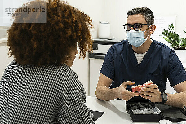 Männlicher Zahnarzt mit Zahnersatz erklärt einer Patientin in einer medizinischen Klinik während der Pandemie