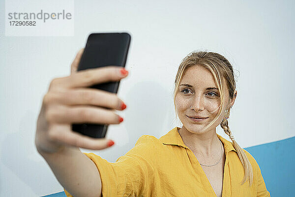 Lächelnde Frau nimmt Selfie durch Handy von weißen Wand