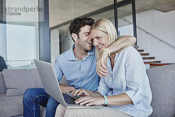 Lächelnder Mann umarmt Frau  die einen Laptop benutzt  während sie auf dem Sofa im Wohnzimmer sitzt