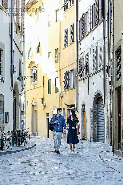 Touristisches Paar mit Gesichtsmaske  das sich beim Gehen auf einer Straße in der Stadt an den Händen hält