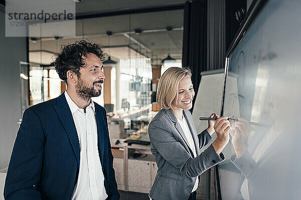 Lächelnde Geschäftsfrau  die auf einer Projektionsfläche schreibt  während sie neben einem Kollegen im Büro steht