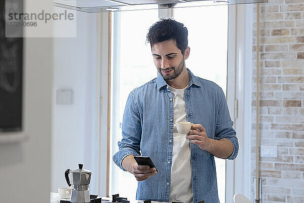 Junger Mann benutzt ein Mobiltelefon und hält eine Kaffeetasse in der Küche