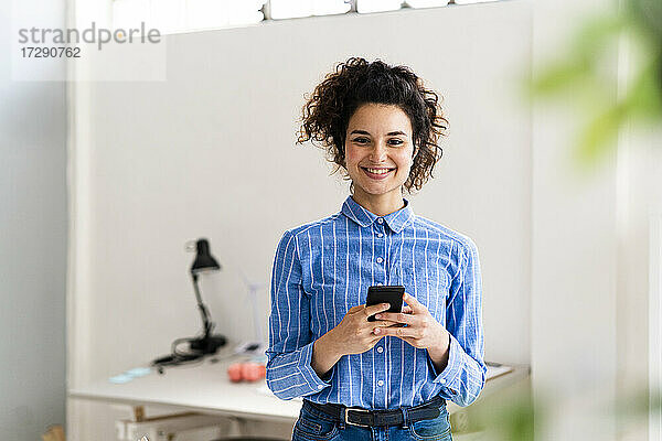 Lächelnde Geschäftsfrau  die ein Smartphone hält  während sie in einem kreativen Büro steht