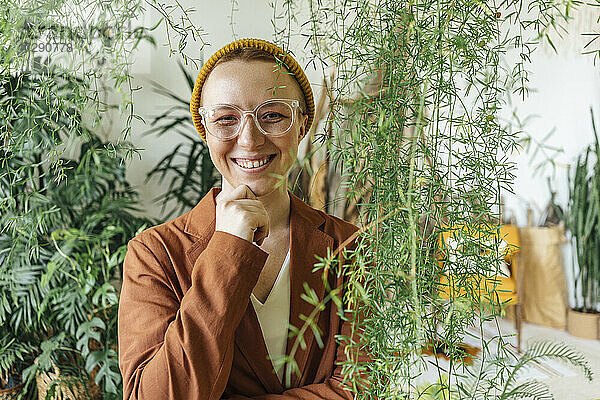 Lächelnde Unternehmerin mit Hand am Kinn  die bei Pflanzen am Arbeitsplatz steht
