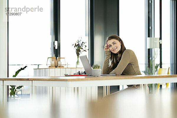 Lächelnde Geschäftsfrau mit dem Kopf in der Hand  die am Tisch im Büro sitzt und nachdenkt