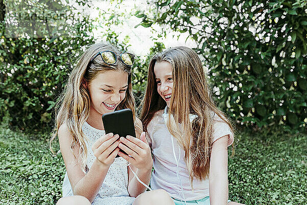 Fröhliches Mädchen mit Freundin  die im Garten ein Mobiltelefon benutzt