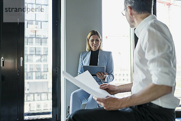 Lächelnde Geschäftsfrau  die ein digitales Tablet benutzt  während sie sich mit einem männlichen Kollegen im Büro unterhält