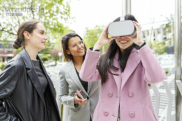 Lächelnde Geschäftsfrau  die ein Virtual-Reality-Headset benutzt  während sie neben ihren Kollegen steht