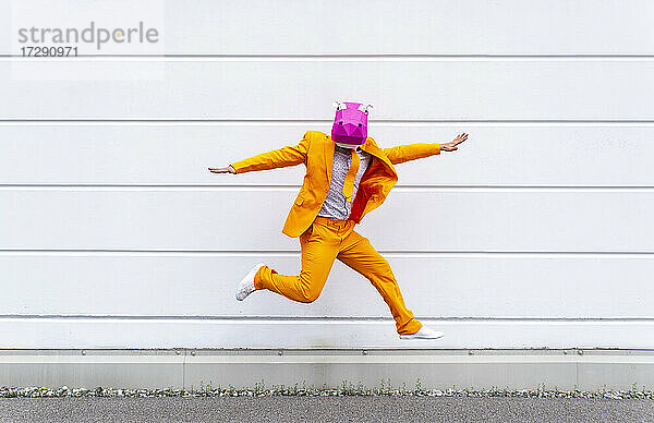 Mann in leuchtend orangefarbenem Anzug und Nilpferdmaske springt vor eine weiße Wand