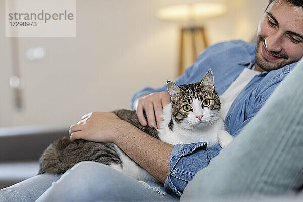 Lächelnder Mann  der seine Katze streichelt  während er zu Hause auf dem Sofa sitzt