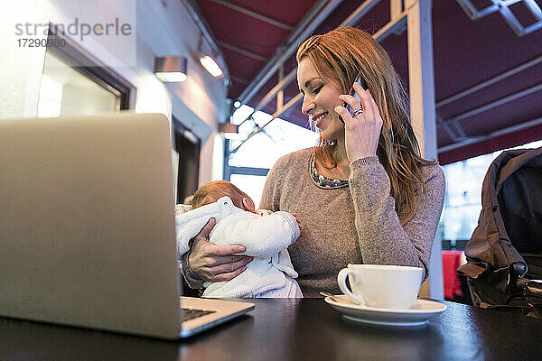 Mutter  die ihr Baby trägt  während sie in einem Café mit dem Handy telefoniert
