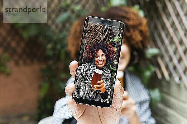 Lächelnde Afro-Frau  die ein Selfie mit ihrem Smartphone macht  während sie Bier trinkt