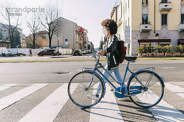 Junge Frau mit Gesichtsschutz beim Überqueren der Straße mit dem Fahrrad an einem sonnigen Tag