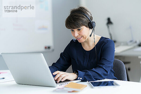 Lächelnde weibliche Fachkraft mit Headset und Laptop am Schreibtisch im Büro sitzend