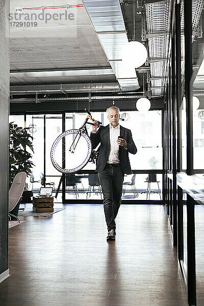 Älterer Geschäftsmann  der ein Mobiltelefon benutzt  während er im Büro ein Fahrrad auf der Schulter trägt