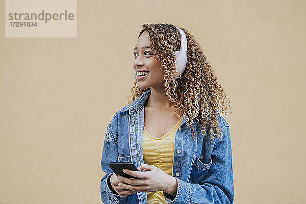 Junge Frau mit Kopfhörern schaut weg und hält ihr Smartphone an einer beigen Wand