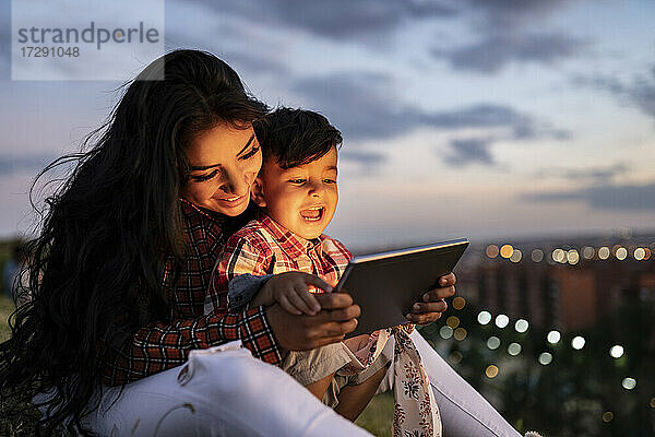 Glücklicher Sohn benutzt digitales Tablet  während er mit seiner Mutter bei Sonnenuntergang sitzt