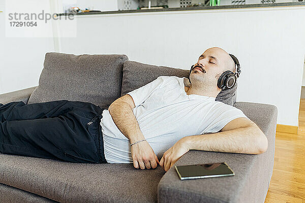 Glatzköpfiger Mann mit geschlossenen Augen hört Musik über Kopfhörer  während er sich zu Hause auf dem Sofa entspannt