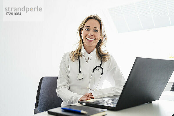 Lächelnde Ärztin mit Stethoskop am Schreibtisch sitzend im Büro