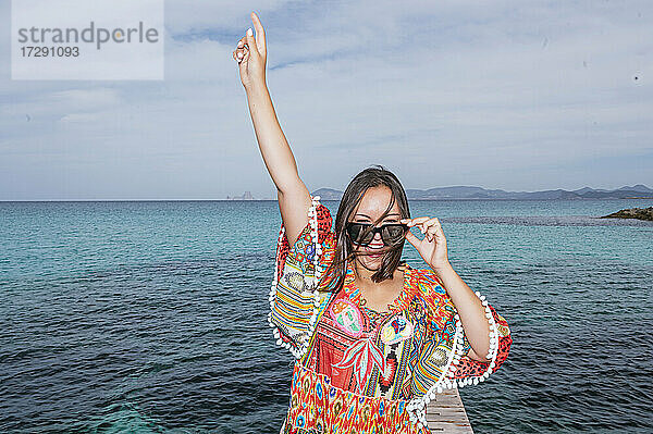 Fröhliche Frau mit erhobener Hand vor dem Wasser auf der Insel Formentera stehend